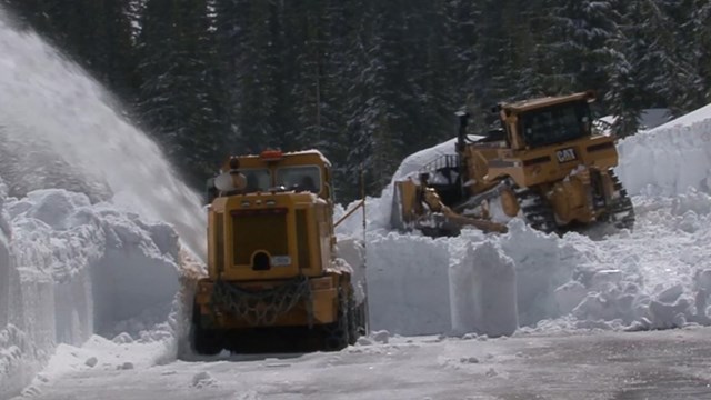 A dozer pushes into deep snow while a rotary snowplow clears another layer to reveal a road surface.