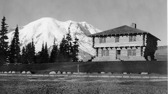 Black and white photo of a log building in front of a view of Mount Rainier. 
