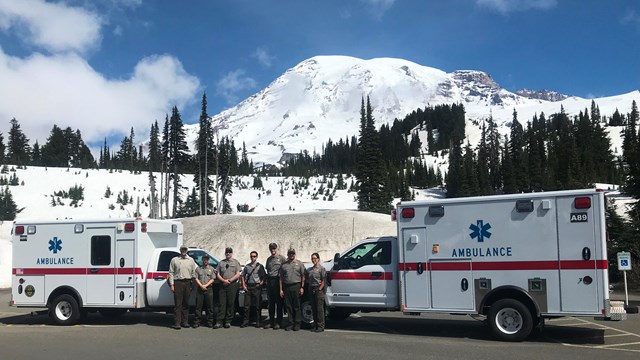 Rangers stand in front of two ambulances posed in front of a snow-covered mountain. 