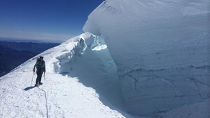 A climber stands on the edge of a large glacier crevasse. 