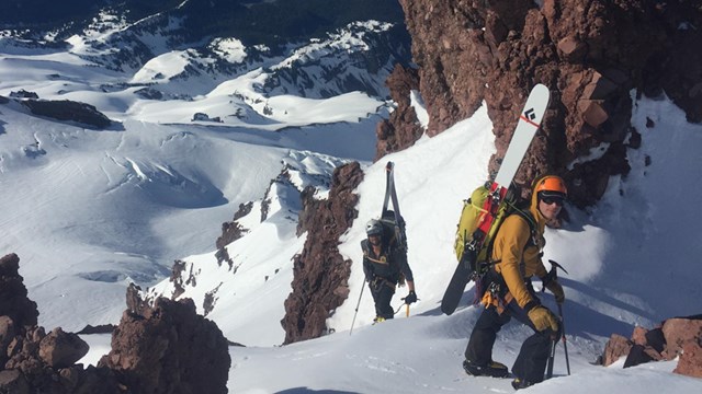Two climbers scale a deep snowy slope high above the surrounding mountain ranges. 
