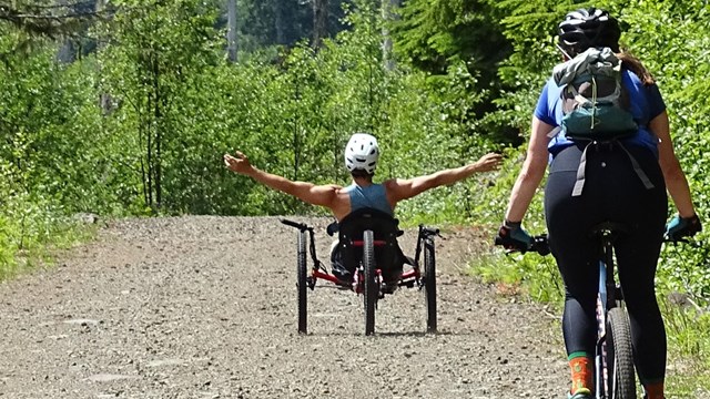 A hand cyclist and mountain biker descend a gravel road.