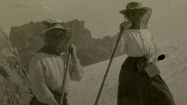 Historic photo of two women holding walking staffs standing on a steep snowy slope. 