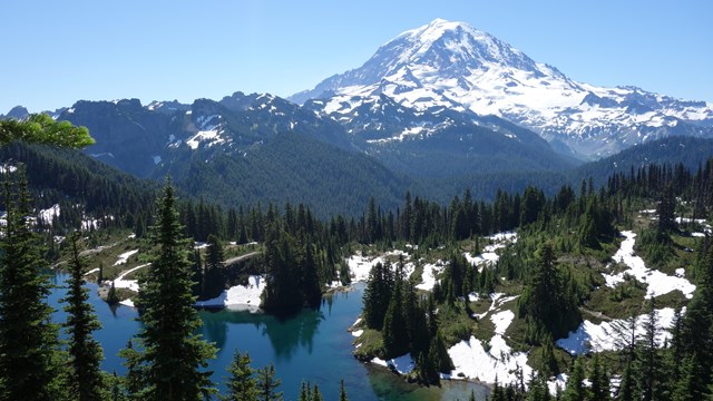 A glacier-covered volcanic peak towers over ridges, forested valleys, and an alpine lake. 