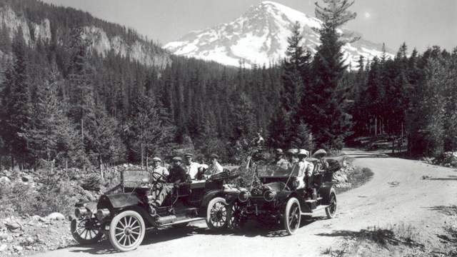 Two old cars on a forested road with Mount Rainier in the background