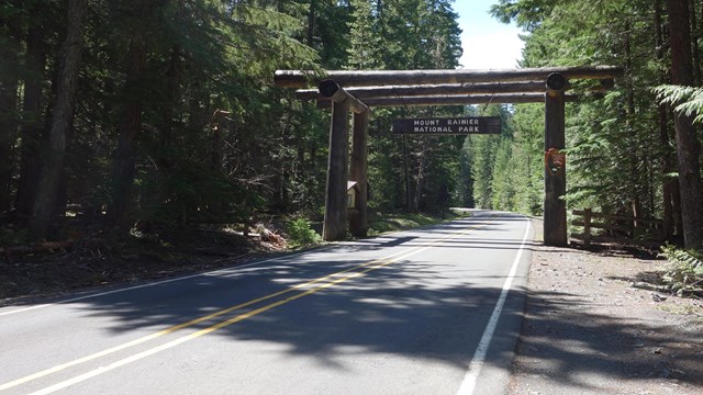 A two-lane paved road goes through a log entrance arch bordered by forest. 