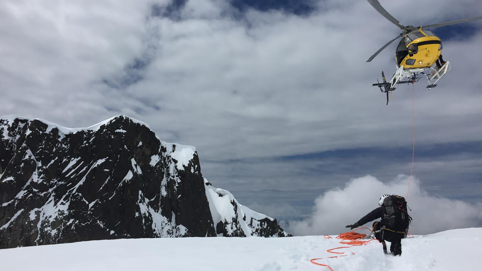 A climber crouches next to a red rope on a snowy slope while a yellow helicopter approaches.