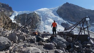 A man stands next to equipment set up on the rocky upper slope of a mountain. 