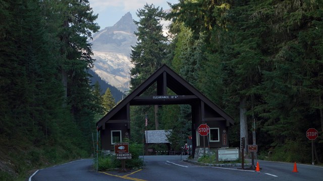 A paved road bordered by forest leads to an entrance with two booths connects by an A-frame roof. 