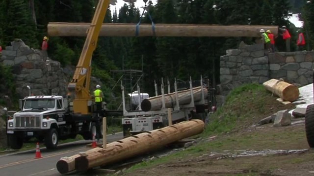 A large truck lifts a large log to span above a road between to rock walls. 