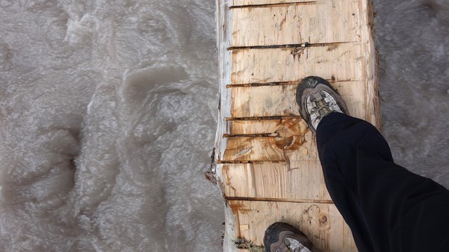 Looking down at a rushing river passing underneath a log footbridge. 