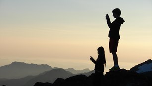 Two children silhouetted against an evening sky and mountain ranges. 