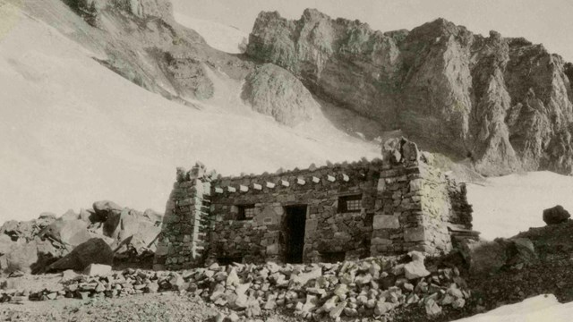 A small rectangular stone building perched on a ridge high on a mountain. 