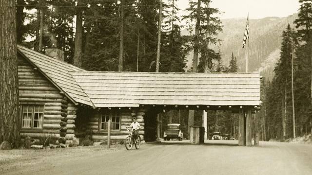 A wood building with a covered porch that extends out over a dirt road. 