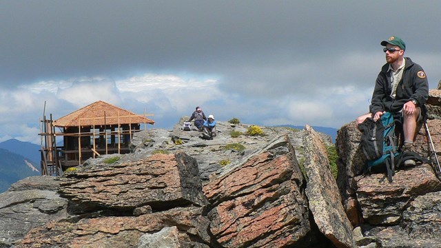 A man wearing a volunteer ranger uniform sits on a rock next to a fire lookout. 