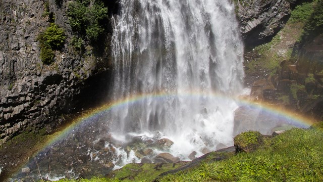 A rainbow arches through the spray of a waterfall cascading down a rocky cliff. 