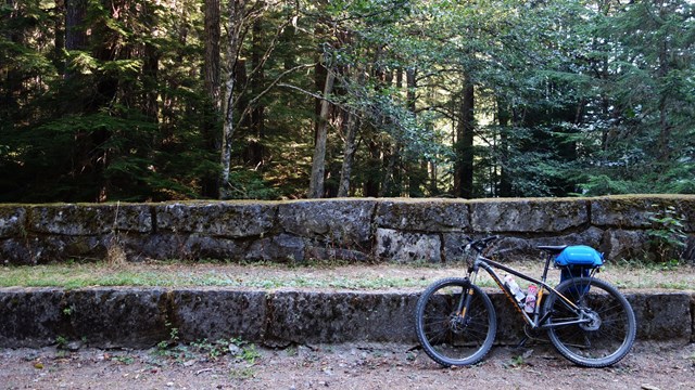 A mountain bike leans against a large stone curb and rock wall along a gravel road. 