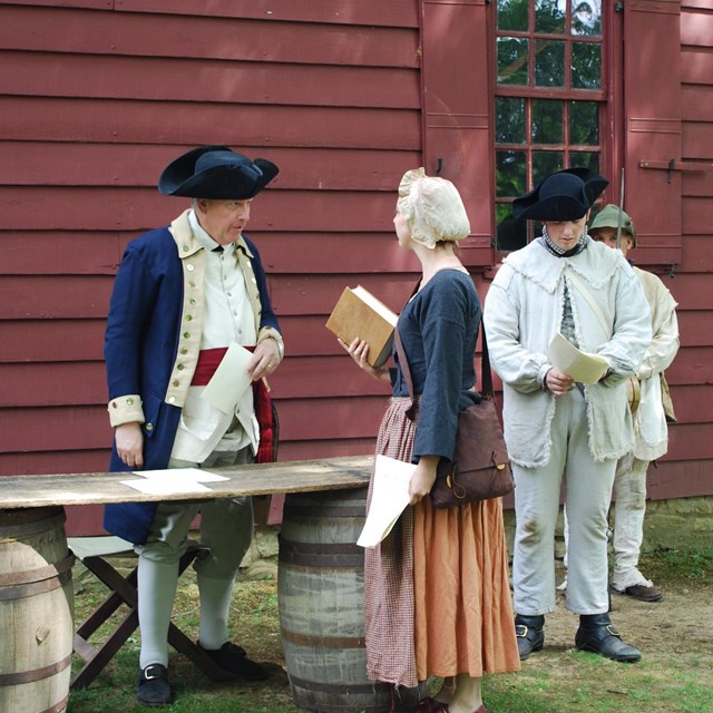A woman addresses a man dressed as an officer as two soldiers look on.