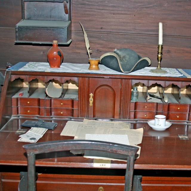 A colonial desk with papers, teacups, a hat, candles, and more on it.