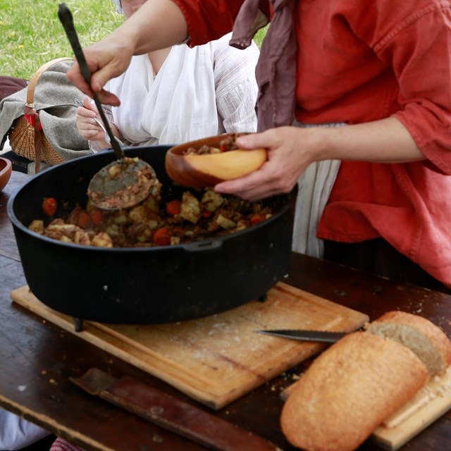 A woman in colonial dress serving stew from a pot at a camp site.