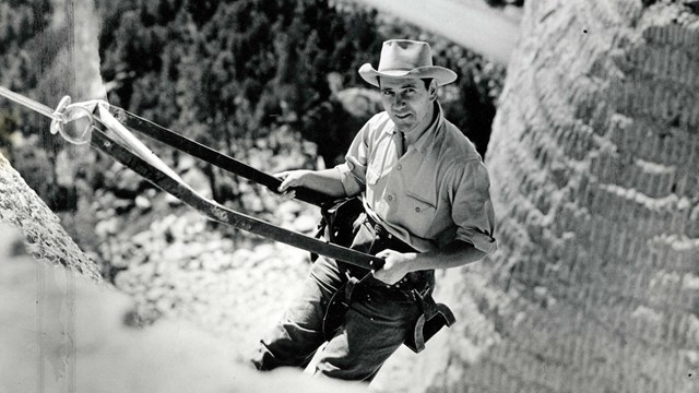 Black and white photograph of Lincoln Borglum in a bosun chair on Mount Rushmore.