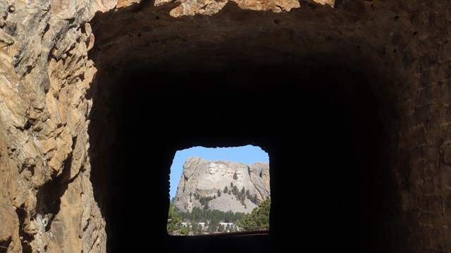Mount Rushmore sculpture is surrounded by a blue sky and is seen at the end of a dark tunnel. 
