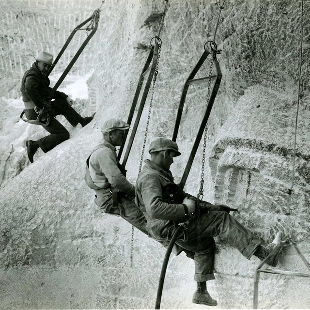 Photo of Gutzon Borglum observing two workers sculpting an eye on Mount Rushmore.