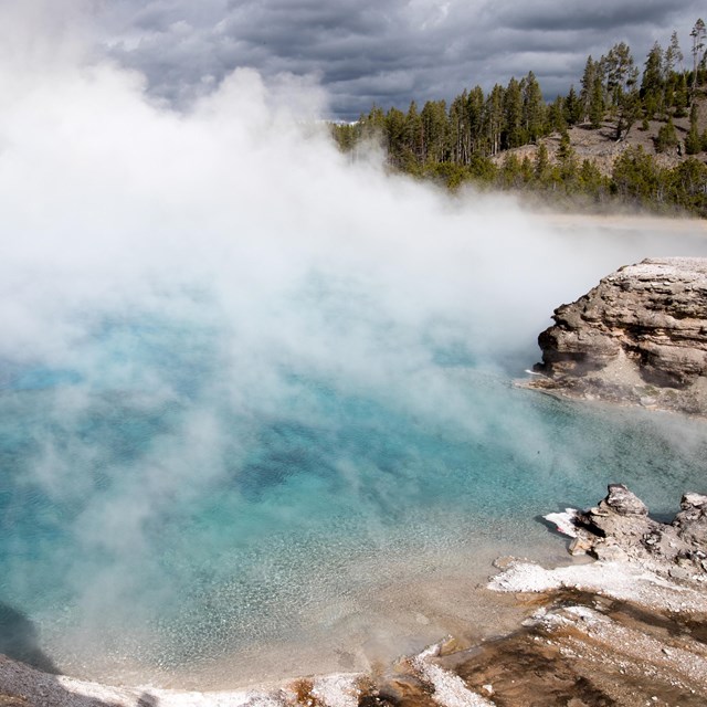 Steam rises off clear blue water surrounded by rocks and forest.