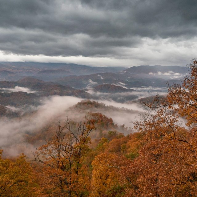 View of red fall color, distant rolling mountains, and grey storm clouds.
