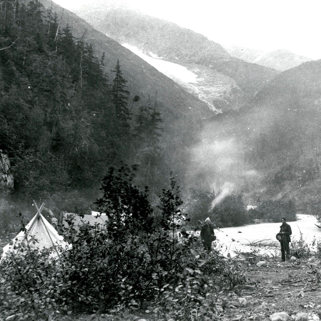 Black and white image of a mountain stream with person on the bank and a tent farther up the bank.