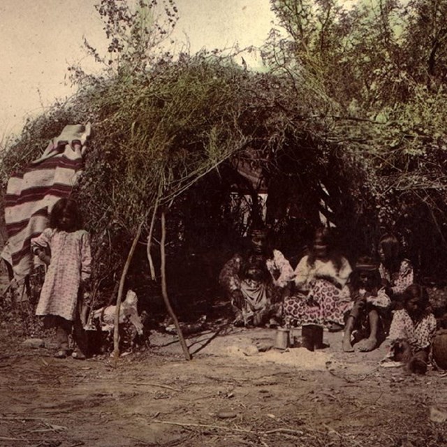 A grainy black and white image of a hut made from sticks (wikiup) with people sitting in front.