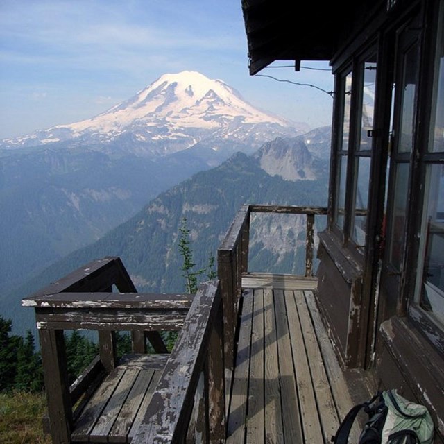 A snow covered mountain in the background seen from a wooden porch on a fire lookout.