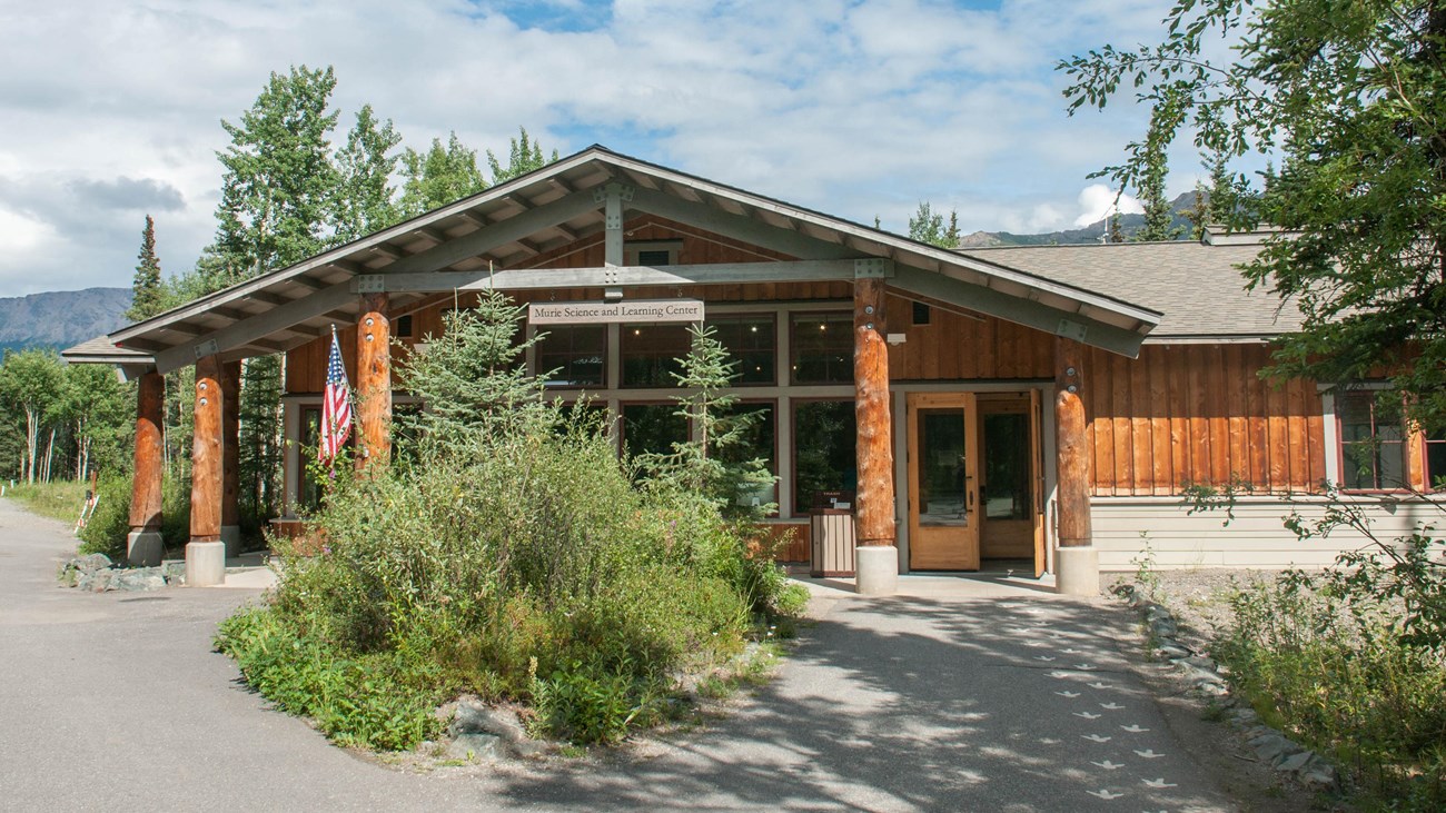 A building with prominently log framing on a partly sunny day. Green vegetation lines the pathway.