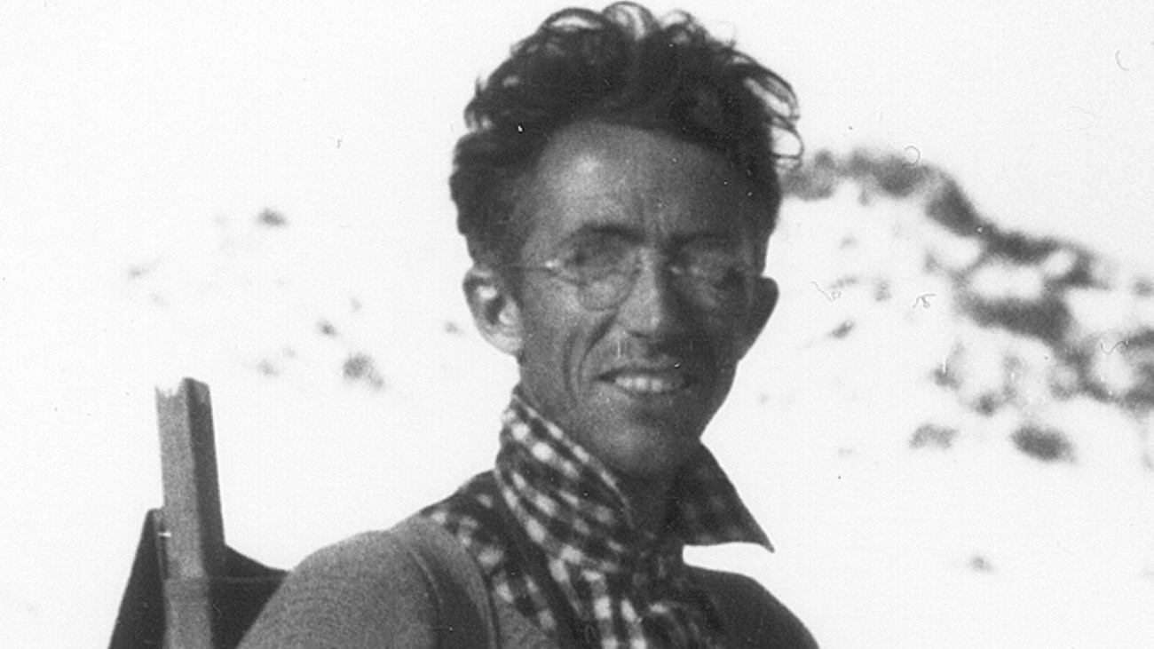 Grainy black and white archival photo of a man standing outside, wearing a backpack and glasses.