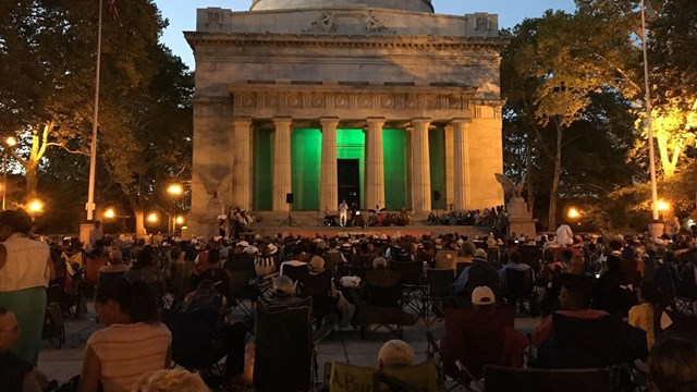 Hundreds of people sit in folding chairs, facing a large white marble sarcophagus