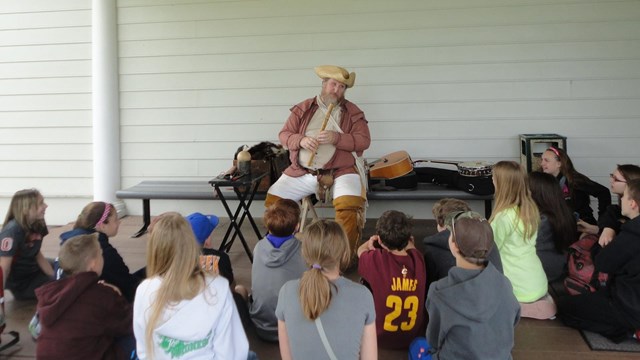 Students sit at the foot of a Man in historic dress hair holding a Fife.