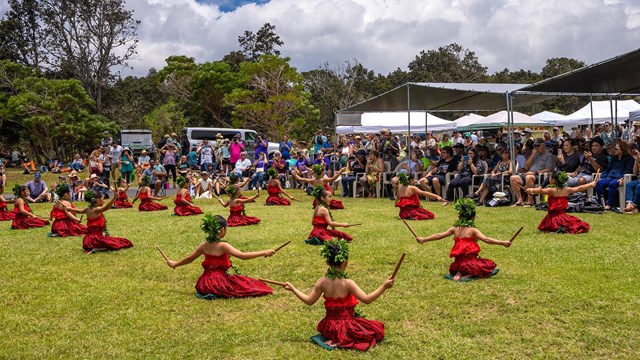 Dancers wearing red dresses and green leaves in their hair play wooden instruments while seate