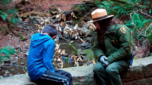 A ranger and a child sit on a bridge and look at the creek