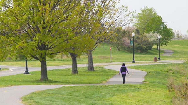 A woman walks on a path surrounded by green trees and grass