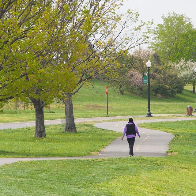 A woman walks on a path surrounded by green trees and grass