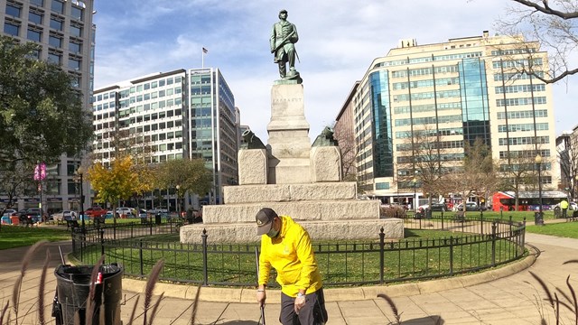 A man in yellow sweeps trash with a statue in the background. 