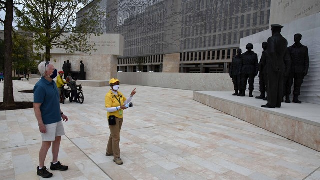 A woman in yellow points to statues with a man in blue and khaki. 