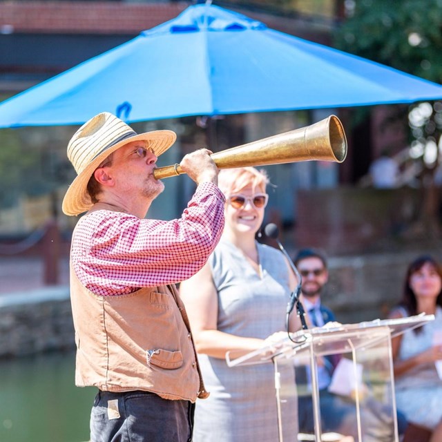 Man plowing a ceremonial horn next to a woman behind a podium