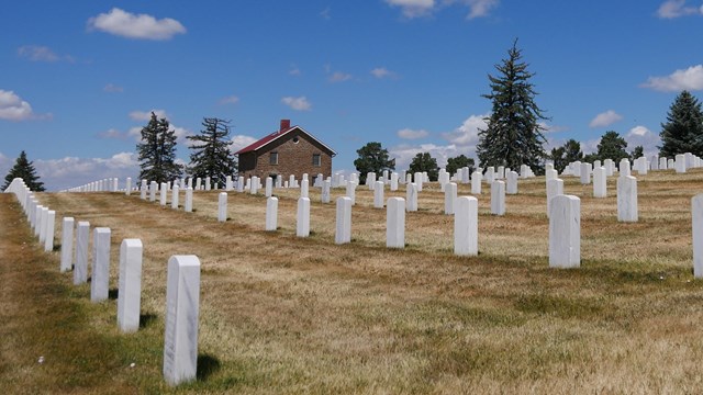 Short, turf around tows of white marble headstones on a slope, with a lodge and pines in the rear.