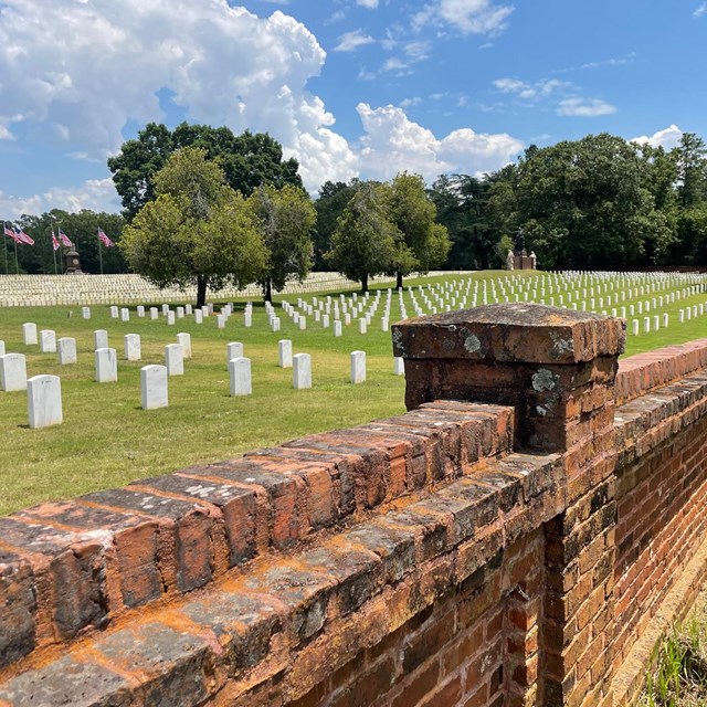 Rows of uniform white marble headstones in cemetery of short turf and trees, within a masonry wall.