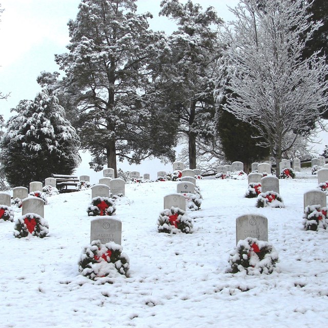 A light snow covers a cemetery scene, where a wreath with red bow rests at the base of each marker.