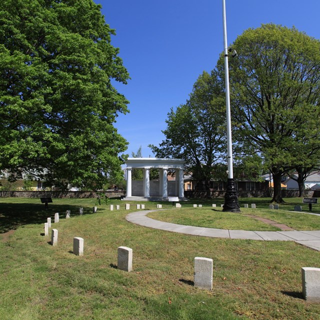 Uniform headstones are arranged in a circle around a walkway and central flagpole on a grassy mound