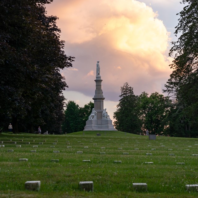 Rows of headstones and turf in front of the Soldiers' National Monument, with a figure on a column