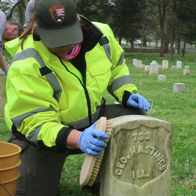An NPS employee uses a brush to clean the surface of a headstone.