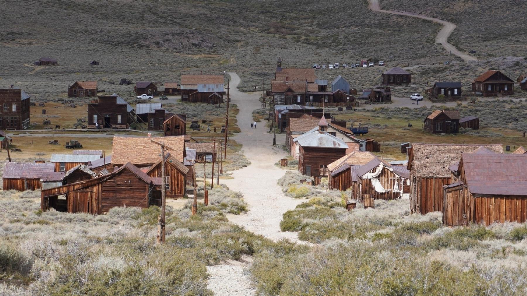 Wood frame buildings lining a dirt road in a desert setting. A road winds up the mountain in the bac